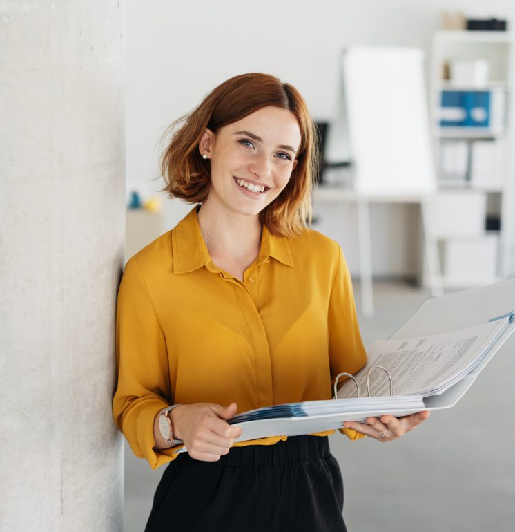 Attractive young office worker holding a large open binder as she looks at the camera with a sweet friendly smile